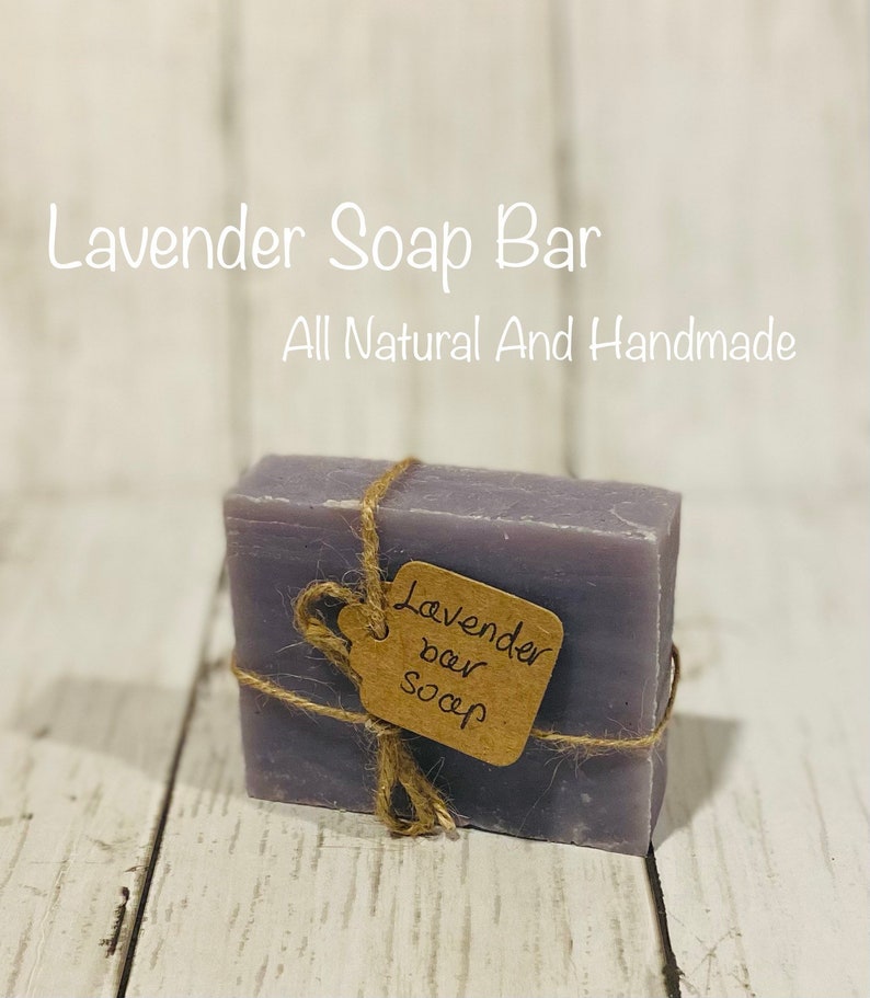 Pu&ograve; includere: Una saponetta di colore lavanda avvolta in spago con un'etichetta di carta marrone su cui &egrave; scritto "Lavender bar soap". La saponetta &egrave; su una superficie di legno bianco. Il testo "Lavender Soap Bar" e "All Natural And Handmade" &egrave; scritto in bianco su uno sfondo bianco sopra la saponetta.