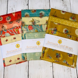 May include: Three sets of beeswax food wraps in various floral patterns. The wraps are in shades of red, teal, and gold. Each set is bundled with a white band that reads "Beeswax Food Wraps" and "My Zero Waste Store".