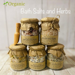 May include: Six glass jars filled with bath salts and herbs, labeled with different scents and ingredients. The jars are arranged in a pyramid shape on a wooden surface. The jars are topped with brown paper and twine.