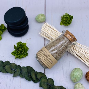 Puede incluir: Un frasco de vidrio lleno de sales de ba&ntilde;o y hierbas etiquetado como "Stress Relief Bath Salts and Herbs" con una tapa de corcho. El frasco est&aacute; sobre una superficie blanca con un manojo de hierba seca y elementos decorativos verdes.