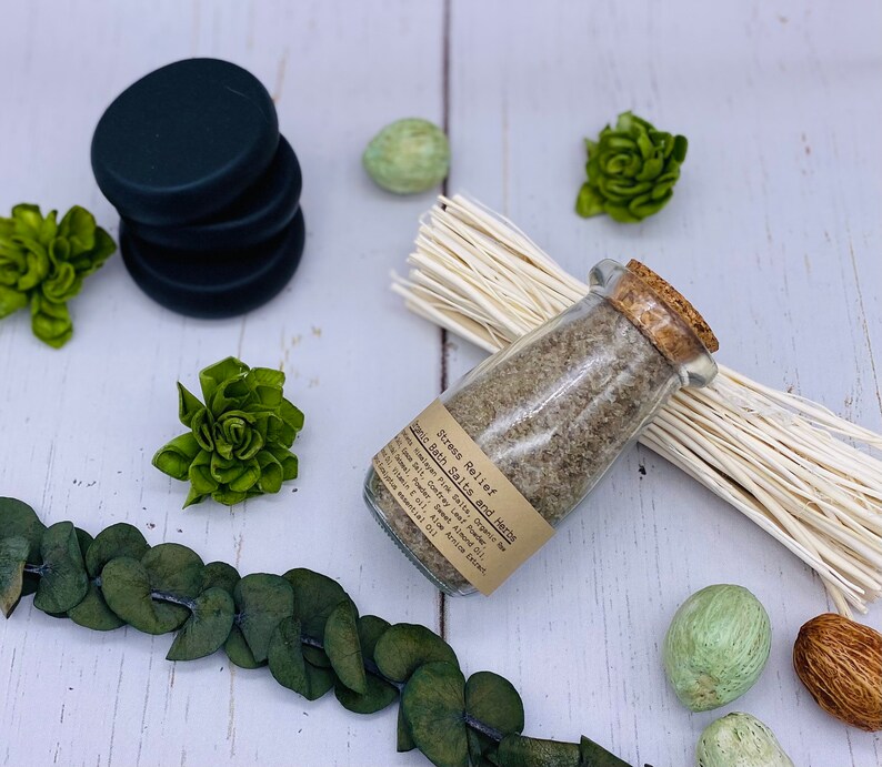 Puede incluir: Un frasco de vidrio lleno de sales de ba&ntilde;o y hierbas etiquetado como "Stress Relief Bath Salts and Herbs" con una tapa de corcho. El frasco est&aacute; sobre una superficie blanca con un manojo de hierba seca y elementos decorativos verdes.