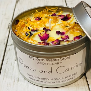 May include: A silver tin candle with a white label that reads "My Zero Waste Store Apothecary Peace and Calming Hand-Poured in Small Batches Made with Love in Pennsylvania". The candle is topped with a white wax layer and decorated with dried yellow petals and pink flower pieces.