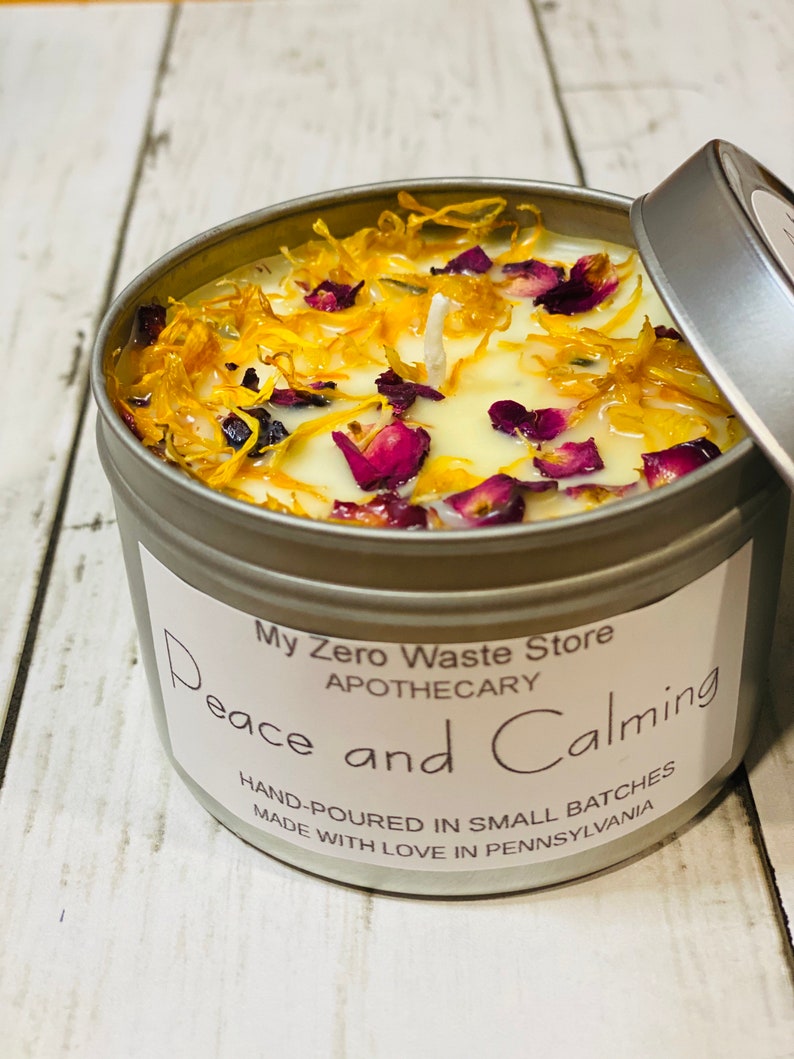 May include: A silver tin candle with a white label that reads "My Zero Waste Store Apothecary Peace and Calming Hand-Poured in Small Batches Made with Love in Pennsylvania". The candle is topped with a white wax layer and decorated with dried yellow petals and pink flower pieces.