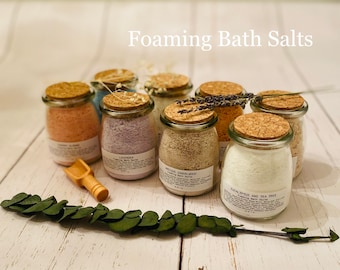 May include: Six glass jars with cork lids filled with different colored bath salts. The jars are labeled with the scents: Lavender, White Tea & Sandalwood, Eucalyptus & Tea Tree, Grapefruit & Rose, and Ginger & Lime. The jars are arranged on a white wooden surface with a sprig of greenery.