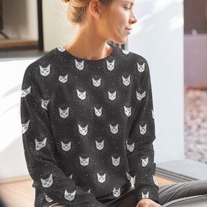 May include: A black sweatshirt with a white cat face pattern. The cat faces have a third eye and a crescent moon above their heads.