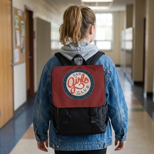 Könnte beinhalten: Ein schwarzer Rucksack mit einer dunkelroten Klappe, die einen runden Aufnäher mit der Aufschrift SAD GIRLS CLUB in weißer und orangefarbener Schreibschrift zeigt. Der Rucksack hat zwei schwarze vertikale Riemen mit Schnallen und wird über einem grauen Kapuzenpullover getragen.