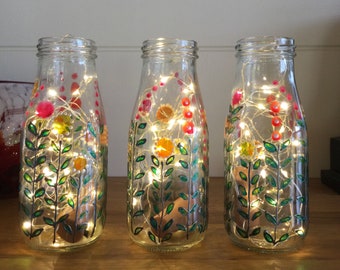 May include: Three glass bottles with painted floral designs and fairy lights inside. The bottles are clear glass and the flowers are painted in shades of green, yellow, orange, and red.