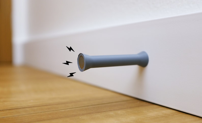 May include: A gray doorstop with a magnetic end, attached to a white door frame. The doorstop is shaped like a cylinder with a rounded end.