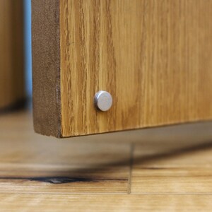 May include: Close-up of a wooden cabinet door with a silver metal hinge. The door is closed and the hinge is visible on the bottom edge of the door.