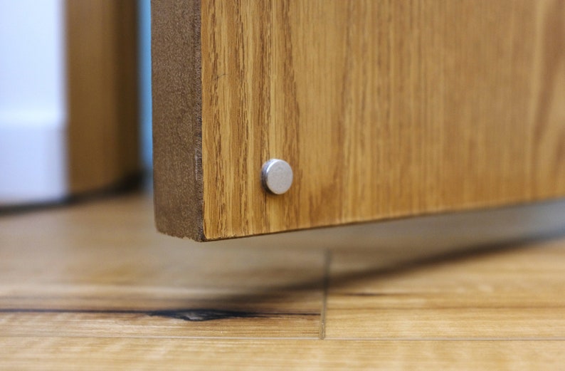 May include: Close-up of a wooden cabinet door with a silver metal hinge. The door is closed and the hinge is visible on the bottom edge of the door.
