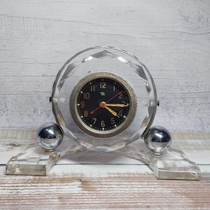May include: A clear, faceted crystal clock with a black clock face and gold-coloured trim. The clock has silver-coloured spherical feet and a clear base. The clock hands are red and gold. The background is a light wood.