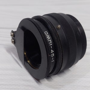 Peut inclure: Un objectif d'appareil photo noir avec une bague en métal et le texte "ОМ-1-45-1" imprimé dessus.