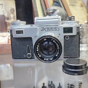Puede incluir: Una cámara de película de 35 mm de color plateado y negro con una lente Helios-103. La cámara tiene una tapa de lente de cuero negro y el texto "Kiev" en la parte superior.