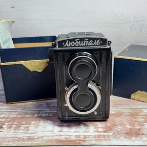 Puede incluir: Una cámara réflex de doble lente negra vintage con la palabra "Любитель" en escritura blanca. La cámara tiene diales plateados y está flanqueada por dos cajas azul oscuro con ribetes dorados, sobre una superficie de madera.