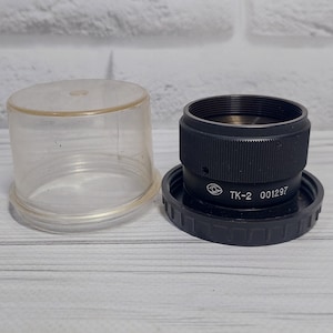 Puede incluir: Un objetivo de cámara negro con una tapa de plástico transparente. El objetivo tiene el texto "TK-2 001297" impreso en él.