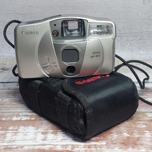 以下が含まれることがあります： 黒いストラップが付いたシルバーのCanon Prima AF-9sカメラが、赤いCanonの文字が入った黒いケースの上に置かれています。 カメラには内蔵フラッシュとさまざまなボタンがあります。 背景は木製の表面です。