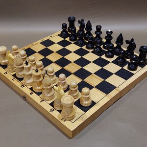 May include: A wooden chessboard with black and white squares. The board is open and shows black and white chess pieces in their starting positions. The board is made of wood and has a light brown finish.
