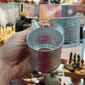 May include: Two silver measuring cups with a star pattern and a handle. The cups are made of metal and have a shiny finish.