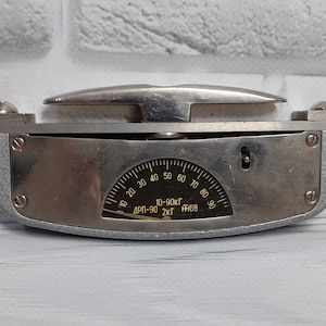 Könnte beinhalten: Ein silberfarbenes, handliches Gerät mit einem Zifferblatt, auf dem "10-90kg" und "ДП-90 2кг" steht. Das Gerät hat einen silberfarbenen Griff und zwei rote Knöpfe an beiden Seiten.