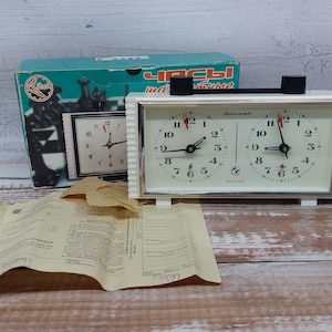 May include: Vintage white chess clock with black accents, featuring two clock faces with black hands and red second hands. A teal box with chess pieces and Cyrillic text is behind the clock, along with a paper instruction sheet.