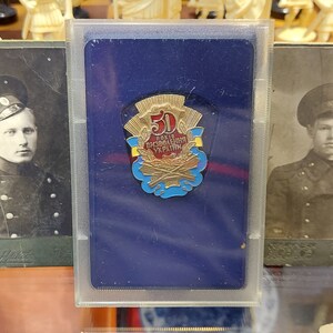 May include: Two vintage black and white photographs of men in military uniforms with hats. The photographs are framed and displayed next to a blue card with a gold pin featuring the text "50 років визволення України" (50 years of liberation of Ukraine).