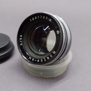 May include: A vintage camera lens with a black and silver finish, resting on a clear plastic container. The lens has visible markings including "2/50" and "10 ИНДУСТАР-6M". A black lens cap sits to the left.