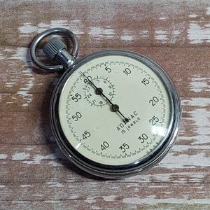 May include: A silver pocket watch with a white face and black markings. The watch has a black hand and a red hand. The watch face reads "ADMAC 1/5 SECONDS".