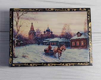 May include: A wooden box with a black and gold painted border. The box features a winter scene with a horse-drawn sleigh, a church, and snow-covered houses. The scene is painted in a realistic style with muted colors.