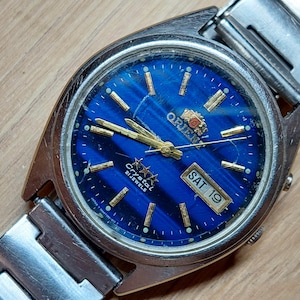 May include: A silver wristwatch with a blue face and gold accents. The watch face has a date window that reads "SAT 19" and the text "Crystal 21 Jewels". The watch brand is "Orient".
