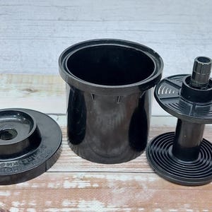 Może przedstawiać: Czarny zestaw do wywoływania filmów, składający się z pokrywy, szpuli i pojemnika, na jasnej powierzchni imitującej drewno. Widoczny jest cylindryczny zbiornik i szpula ze spiralnym wzorem.