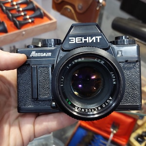May include: A black Zenit Automat film camera with a black lens labelled "AUTO REVUE NON MC 1:1.7 f=50mm JAPAN".