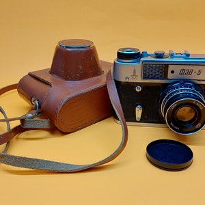 May include: A silver and black 35mm film camera with a leather case and a lens cap. The camera has a silver body with black accents and a black lens. The case is brown leather with a strap. The lens cap is black plastic.