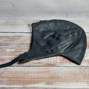 May include: A dark blue leather aviator helmet with a strap and button closure. The helmet has a textured surface and a soft, brown interior lining. The design includes a small pocket and a strap with a buckle.
