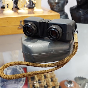 May include: A black and gray vintage viewer with a brown cord and a gray case. The viewer has two round lenses and a ribbed design.