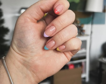 Puede incluir: Primer plano de una mano con uñas ovaladas pintadas con una base nude y puntas de colores. Las puntas están pintadas con un patrón de arcoíris, con cada uña con un color diferente: rosa, morado, azul, amarillo y naranja.
