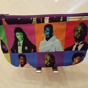 May include: A purple zippered pouch with a colorful pop art design featuring multiple portraits of people. The pouch is made of fabric and has a zipper closure.