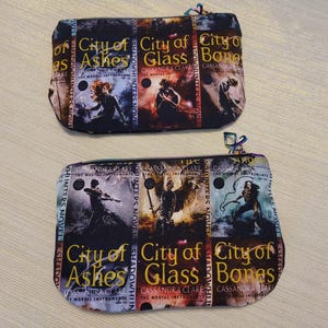 May include: Two zippered pouches with a book cover design. The pouches feature the titles "City of Ashes", "City of Glass", and "City of Bones" by Cassandra Clare. The design includes images of characters and the series title.