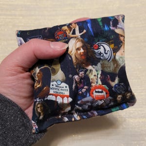 May include: A small, black fabric pouch with a collage of images from the TV show "The Vampire Diaries". The images include characters, symbols, and locations from the show. The pouch is filled with small, soft objects, likely for use as a stress ball or fidget toy.