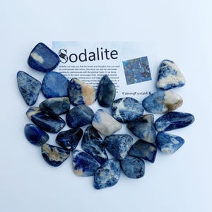 May include: A collection of tumbled sodalite stones. The stones are a deep blue with white veins and are arranged in a loose pile. The image includes a card with the text "Sodalite" and information about the stone's properties.