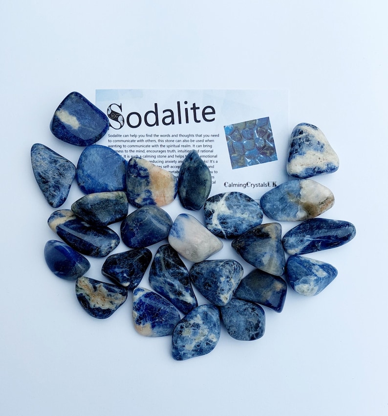 May include: A collection of tumbled sodalite stones. The stones are a deep blue with white veins and are arranged in a loose pile. The image includes a card with the text "Sodalite" and information about the stone's properties.