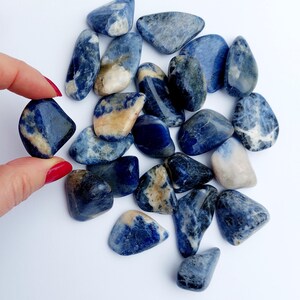May include: A collection of tumbled blue sodalite stones. The stones are various shades of blue with white and brown markings. The stones are smooth and polished.