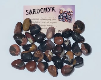 May include: A collection of approximately 30 smooth, polished, dark brown, red, and black sardonyx stones. The stones are arranged in a pile on a white surface. The image includes a small card with the text "SARDONYX" and a description of the stone's properties.