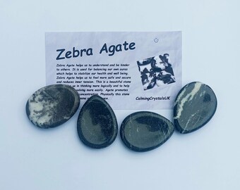 May include: Four black and white zebra agate stones, each with a unique pattern. The stones are smooth and polished, and they are arranged in a row on a white background. The text "Zebra Agate" and a description of the stone's properties are visible on a card behind the stones. The card also includes the text "CalmingCrystalsUK".