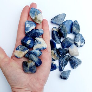 May include: A collection of tumbled blue and white sodalite stones. The stones are smooth and polished, with a variety of shapes and sizes.