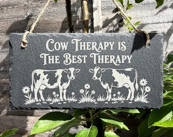 May include: A rectangular dark gray slate hanging sign with a rough edge, featuring the engraved white text "COW THERAPY IS THE BEST THERAPY" above two cows standing in a field of flowers, suspended by a piece of natural jute twine rope.