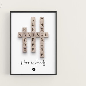 May include: A framed wall art print featuring wooden letter tiles arranged in a crossword style to spell the names Kyde, Madison, Juniper, and Theodore. Below the names, the text reads Home is Family in black script, followed by two small hearts.