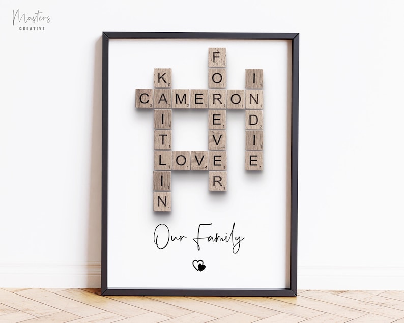 May include: A framed wall print featuring wooden letter tiles arranged in a crossword style to spell out the names Kaitlin, Cameron, and Indie, along with the words Forever and Love. Below the tiles, the text Our Family is printed with a heart icon.