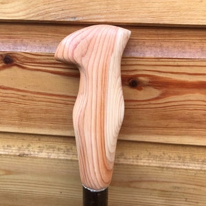 May include: A light brown wooden walking stick handle with an ergonomic shape. The handle, showing visible wood grain, is attached to a dark brown metal shaft. The design suggests comfort and support for the user.