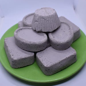 Può includere: Una collezione di oggetti grigi e porosi simili a sapone, di varie forme, tra cui una piccola forma a cupcake, disposti su un piatto verde brillante. Gli oggetti sono impilati e disposti per riempire il piatto.