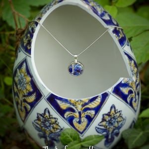 May include: A silver necklace with a blue and white patterned pendant hangs inside a white ceramic egg with blue and yellow floral designs. The egg has a textured surface and is decorated with a repeating pattern.